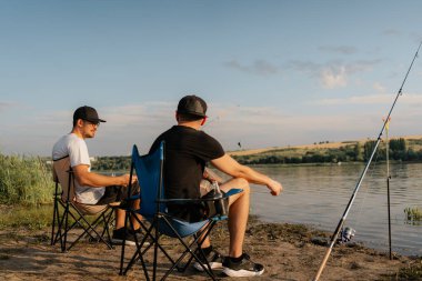 İki adam katlanan sandalyelerde oturup sakin bir gölün kıyısında altın saatinde balık tutarak, rahatlatıcı bir yaz öğleden sonranın tadını çıkarıyor..