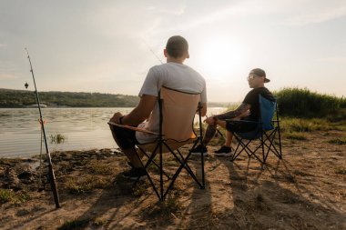 İki arkadaş, gün batımında sakin bir gölün kenarında katlanan sandalyelerde oturup rahatlama ve sohbetin tadını çıkarıyorlar..