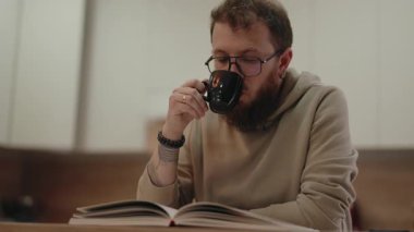 Gözlüklü sakin sakallı adam kahve içer ve evde huzurlu bir anı kucaklayarak kitap okur..