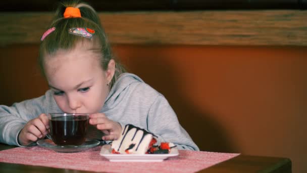 Το κοριτσάκι τρώει επιδόρπιο με φρέσκες φράουλες και πίνει τσάι. Το παιδί χαμογελάει γλυκά ενώ τρώει. Το μωρό φέρνει μια φρέσκια φράουλα στο στόμα της.. — Αρχείο Βίντεο