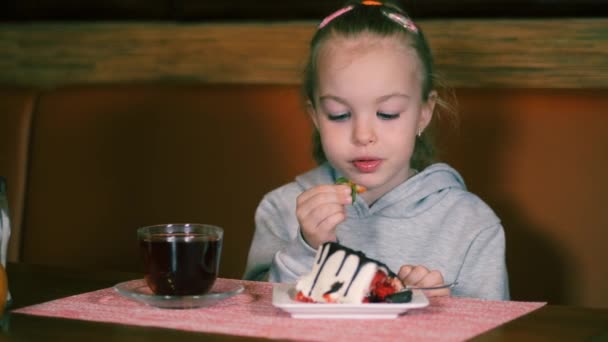 Liten flicka äter dessert med färska jordgubbar och dricker te. Barn ler sött medan de äter. Barnet ger en färsk jordgubbe till sin mun. — Stockvideo