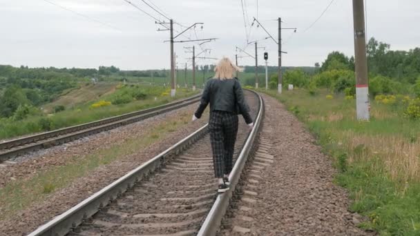 Young lonely woman walks along railroad track balancing on a rail. View from behind. — Stock Video