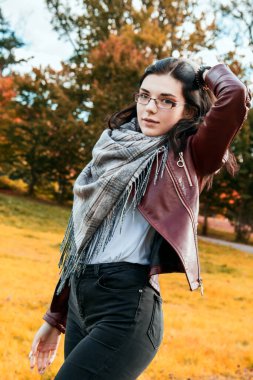 Kahverengi ceketli ve siyah kotlu genç kız güneşli bir sonbahar gününde şehir parkında duruyor.