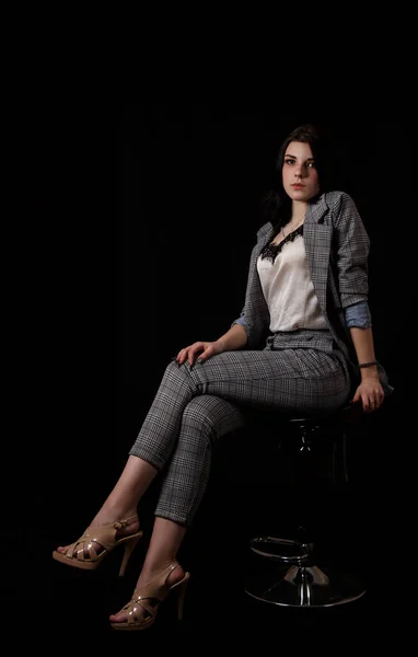身穿西装的年轻貌美的姑娘穿着黑色背景的工作服坐在工作室的椅子上 — 图库照片