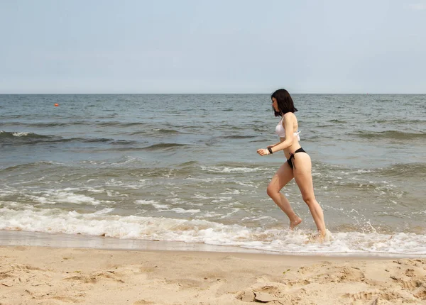 夏の晴れた日に海岸の砂の上を走る黒い水着の若い美しいブルネットの女の子 — ストック写真