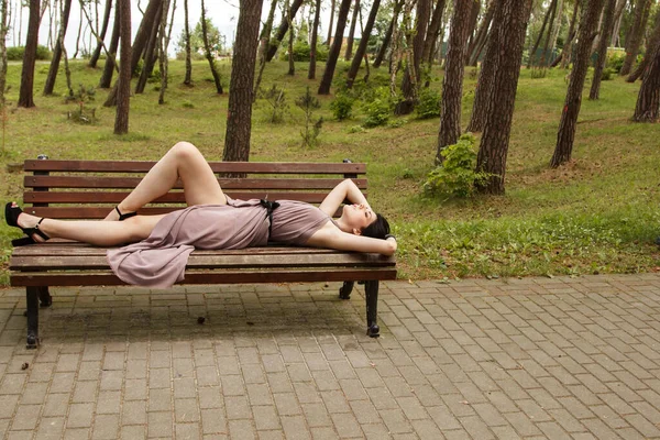 夏の日に公園のベンチに横たわっているベージュのドレスの若い美しいブルネットの女性 — ストック写真