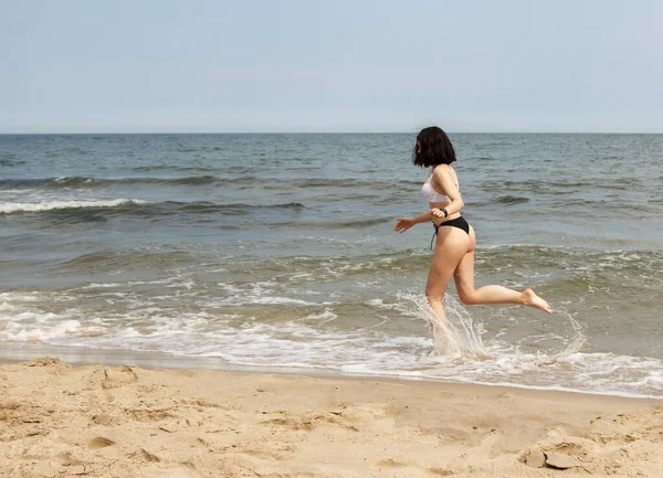 夏の晴れた日に海岸の砂の上を走る黒い水着の若い美しいブルネットの女の子 — ストック写真
