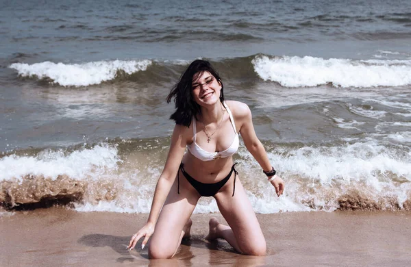 若いですブルネット女性でビキニ姿日当たりの良い夏の日に海によって砂の上にひざまずいて — ストック写真