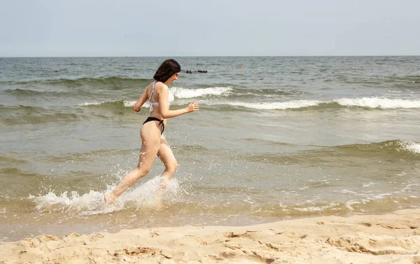 夏の晴れた日に海岸の砂の上を走る黒い水着の若い美しいブルネットの女の子 — ストック写真