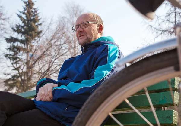 Eldely man sitter på en bänk nära hans cykel i en park — Stockfoto