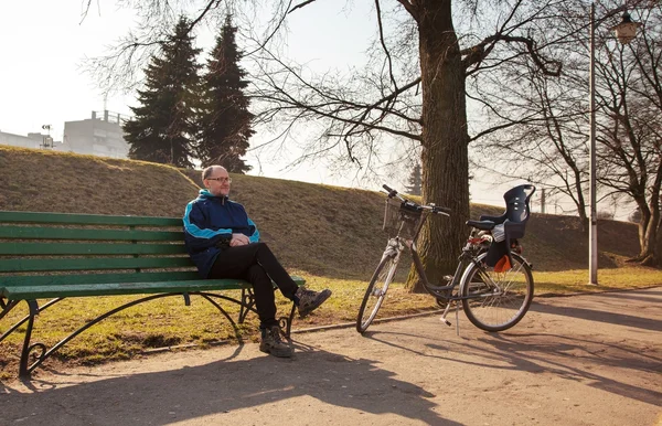 Anziano seduto su una panchina vicino alla bicicletta in un parco cittadino — Foto Stock