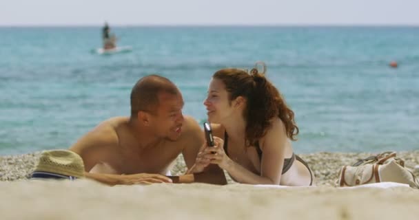 Pareja relajándose en la playa con un teléfono celular — Vídeos de Stock