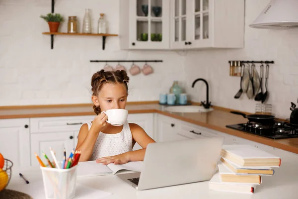 一个漂亮的小女孩 两个马尾辫坐在厨房的桌子旁 手里拿着练习本和笔记本电脑 一边喝茶 一边在学校上课 因为她被隔离了 家庭家庭教育概念 图库照片