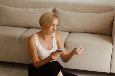 Genç, güzel, sarışın bir kadın akıllı telefon kullanarak oturma odasında oturmuş kredi kartıyla alışveriş yapıyor.