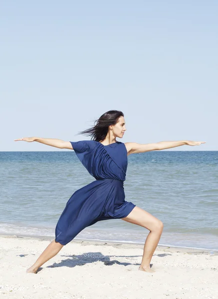 Young beautiful woman doing yoga at seaside in blue dress — Stock Photo, Image