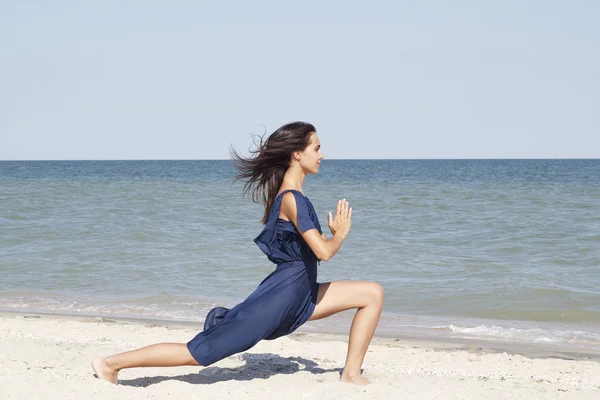 Young beautiful woman doing yoga at seaside in blue dress — Stock Photo, Image