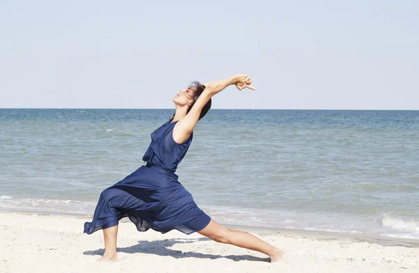 Young beautiful woman doing yoga at seaside in blue dress — Stock Photo, Image