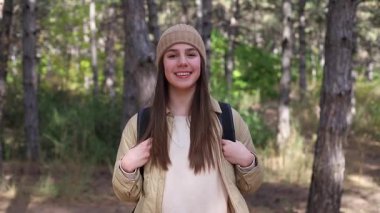 Rahat kıyafetli genç bir kadın ormanda duruyor, sırt çantasını düzeltirken içten bir gülümseme, neşeli bir açık hava anı sergiliyor, kamera yavaşça yaklaşıyor.
