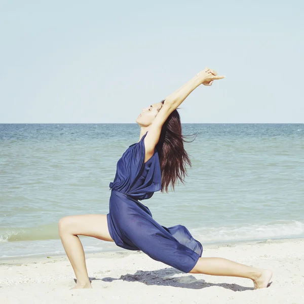 Young beautiful woman doing yoga at seaside in blue dress — Stock Photo, Image