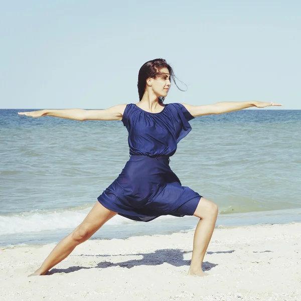 Young beautiful woman doing yoga at seaside in blue dress — Stock Photo, Image