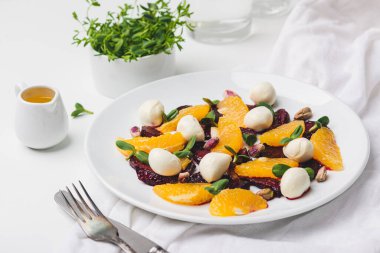 Beyaz bir masada beyaz bir tabakta, sulu meyvelerden oluşan sağlıklı bir salata, lezzetli sebzeler, tuzlu peynir ve çıtır fındıklar.