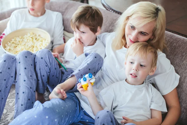 mother and sons are sitting on the couch watching television with popcorn in the kitchen