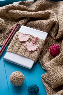 One old notebook in knitted cover with felt hearts lie next to the coil bright filaments and blanket knitted on blue background clipart