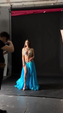 Bir model mavi bir elbisenin içinde güvenle dururken bir fotoğrafçı onun pozlarını karanlık bir arkaplanda çekiyor. Işıklar çalıştıkça ayarı aydınlatır.