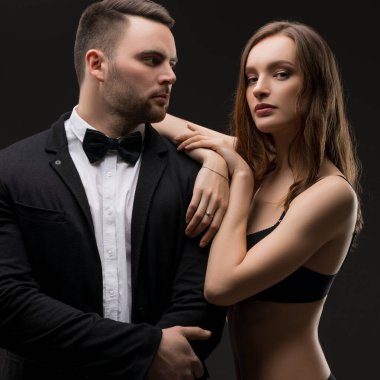 Resmi giyinmiş bir adam ve iç çamaşırlı bir kadın, özgüven ve zarafet sergiliyorlar. Tarzlarını karanlık bir arka planda sergiliyorlar..