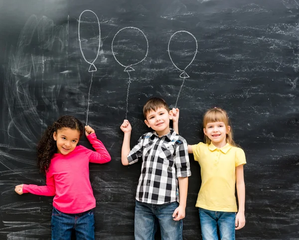 Tres niños alegres mantienen globos imaginarios dibujados en la pizarra —  Fotos de Stock