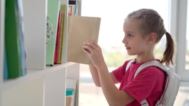 Liten jente søker i bok på bibliotekshylla – stockvideo