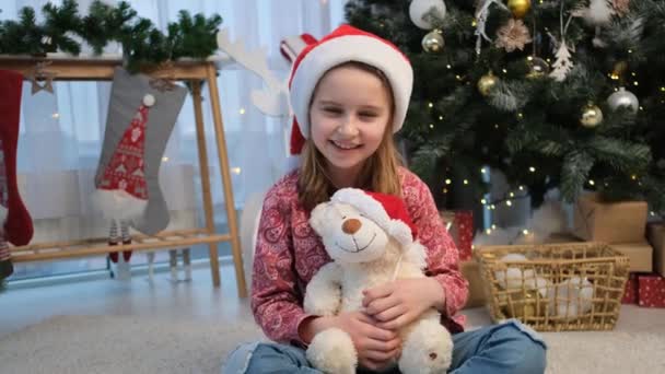 Маленька дівчинка в капелюсі Санта обіймає плюшевого ведмедя — стокове відео