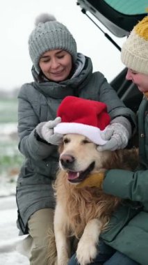 Mutlu bir anne ve kızı, bir araba bagajında bir köpekle kış tatilinde altın renkli, dikey bir videoda Noel Baba şapkası takmayı bırakıyorlar.
