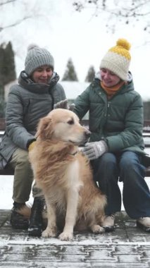 Mutlu küçük kız annesiyle yürüyor ve kış boyunca parkta sevimli bir köpek okşuyor, dikey video.