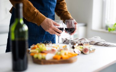 Önlüklü adam şarap kadehlerini hazırlıyor ve iki kişilik romantik bir akşam yemeği için peynir tahtası hazırlıyor.