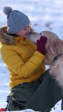 Mutlu bir kadın kışın karlı bir çayırda sevimli bir köpekle yürüyor ve okşuyor.