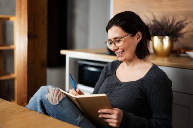 Mutfakta not defterine notlar yazan mutlu genç bir kadın.