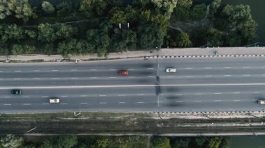 Yukarıdan bakıldığında, yeşil ağaçlarla çevrili ve nehrin yanındaki yolda giden araçlar görülüyor.