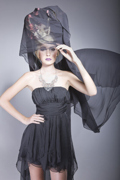 Model posing in a short black dress and a long black veil, attached to a fashionable hat