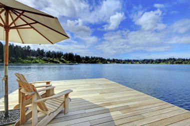 Rıhtımda mavi göle bakan şemsiyeli iki adirondack ahşap sandalye.