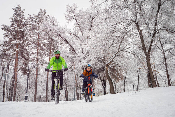 Father and son ride bicycles in the winter park. Bearded man and boy on bicycles spend family weekend in beautiful snowy winter park.