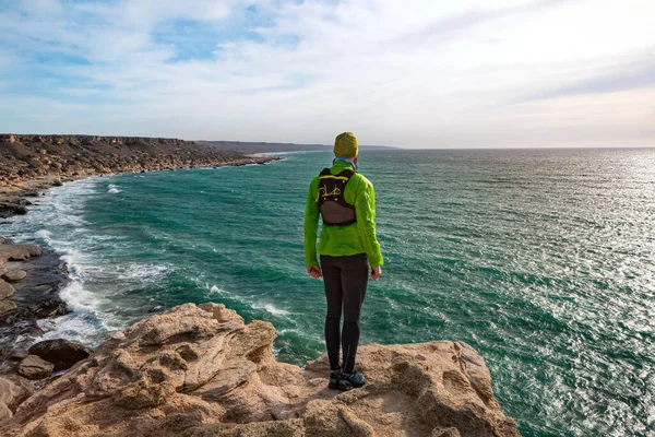 Trailrunner guy stands on the edge of a cliff by the sea on a sunny day. View from the back. Mangystau Peninsula. Kazakhstan