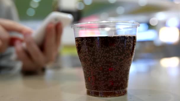 Eine Tasse Coca Cola auf dem Tisch mit einer Frau, die eine Nachricht auf dem iPhone am Food Court im Einkaufszentrum vorliest — Stockvideo