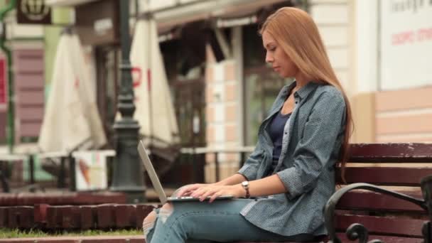 Joven mujer con estilo con un ordenador portátil está sentado en un banco y trabajando . — Vídeo de stock