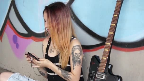 Fille avec tatouage assis près du mur de graffiti avec une musique d'écoute de guitare . — Video