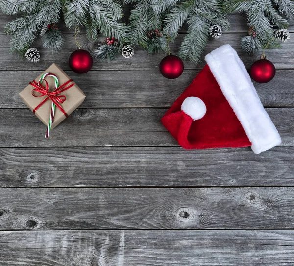 雪に覆われた常緑樹の枝明るい赤いボールの装飾品 ギフトボックス サンタの帽子陽気なクリスマスや幸せな新年のお祝いのコンセプトのための色あせた木製の板 — ストック写真
