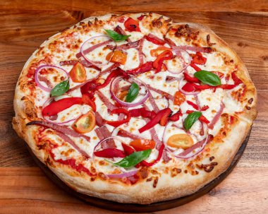 Mozzarella, fesleğen, kırmızı soğan ve biberli yuvarlak pizza. Mozzarella peynirli sıcak yuvarlak pizza, kırmızı soğan halkaları, kırmızı dolma biber ve ahşap masadaki fesleğen yaprakları..