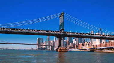 Doğu Nehri ve şehir merkezindeki ufuk çizgisinde Manhattan Köprüsü 'nün üzerinde büyük bir kule var. Suyun düşük açılı görüntüsü, parlak mavi gökyüzüne karşı koyu çelik ve taş yapının ölçeğini vurguluyor..