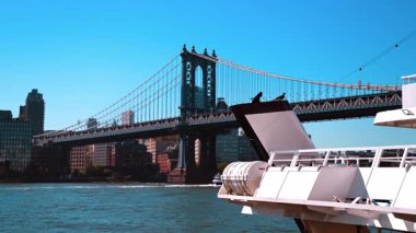 Beyaz bir feribotun ön kısmı Manhattan Köprüsü 'nün çelik yapısını çerçeveler. Yan bakış açısı, asma köprünün endüstriyel yapısını vurguluyor. Doğu Nehri 'nin üzerindeki açık mavi gökyüzünün altında..