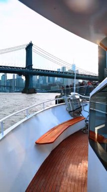New York, ABD, 1 Kasım 2025: Günbatımında Manhattan Köprüsü ve şehir merkezindeki bir tekne güvertesinden görüntü. Geminin ahşap güvertesini ve karanlık köprü yapısı ve şehir manzarasına karşı beyaz korkulukları vurgulayan dikey yakın bir perspektif.